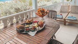 A wooden table with a variety of snacks and a fruit bowl, set on a balcony with a view of a beach and ocean. - Hotel La Plantation (Orient Bay)