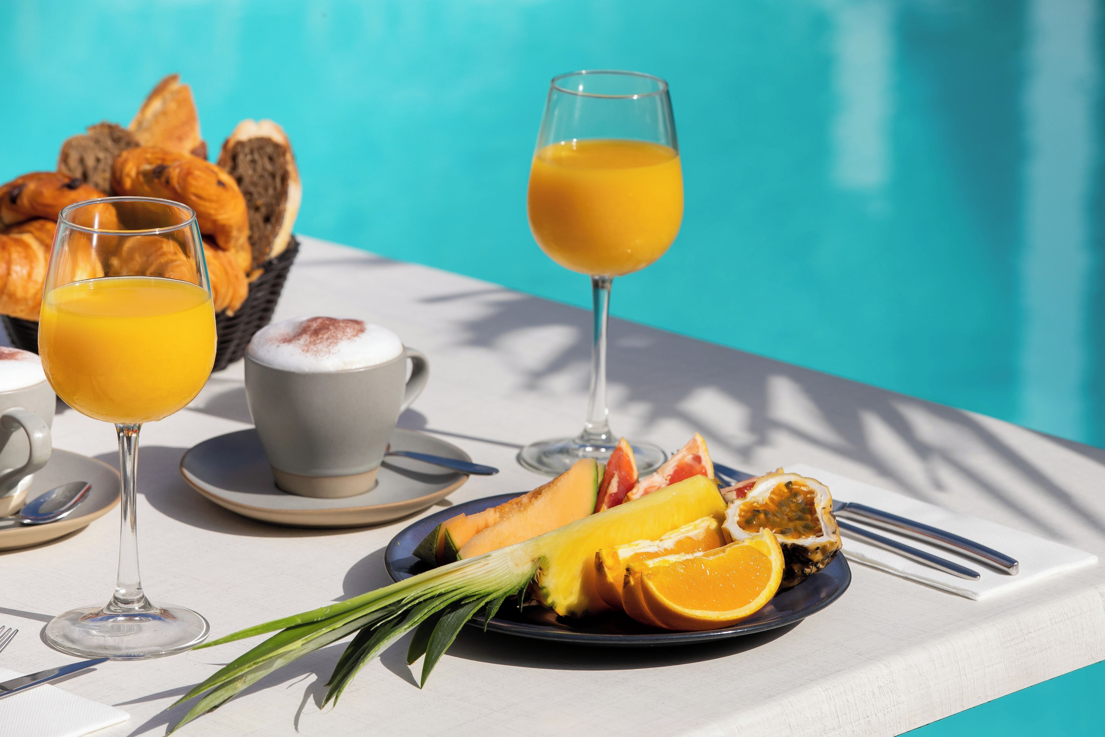 A breakfast setting with croissants, orange juice, a cup of coffee, and a plate of fruit by a poolside.