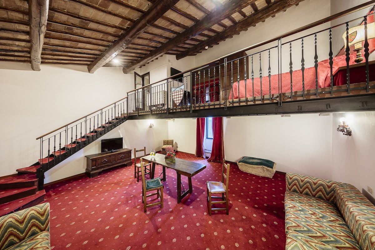 Una habitación con escalera de madera, una mesa de comedor con sillas, un sofá y una alfombra estampada.