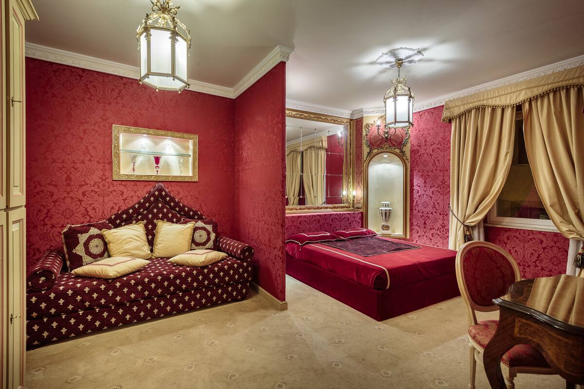 Una habitación con un muro rojo estampado, un espejo grande, una cama con colcha roja, un sofá con funda estampada y una silla de madera.