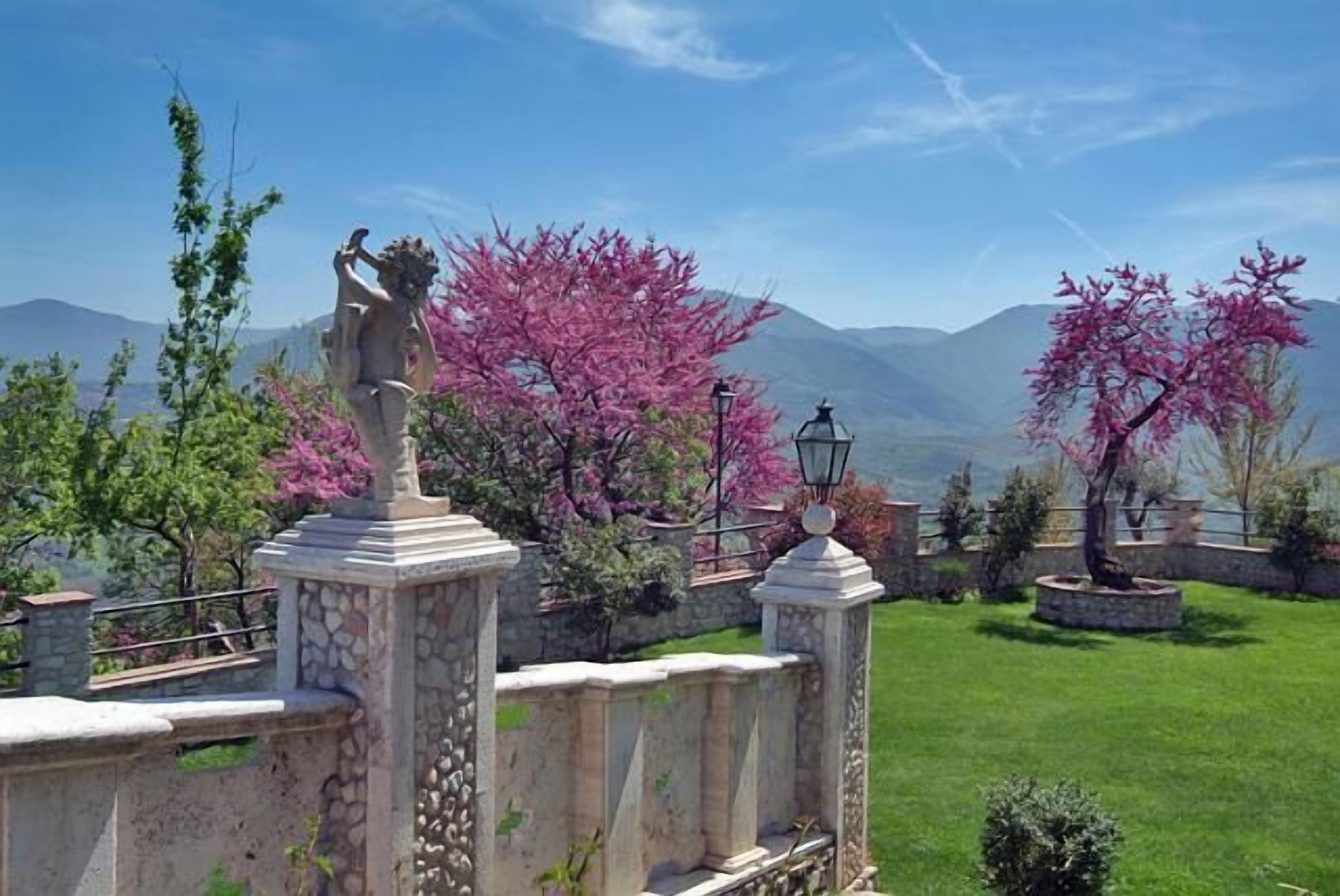 Una balaustra in pietra con una statua, circondata da alberi in fiore rosa e un paesaggio montuoso.