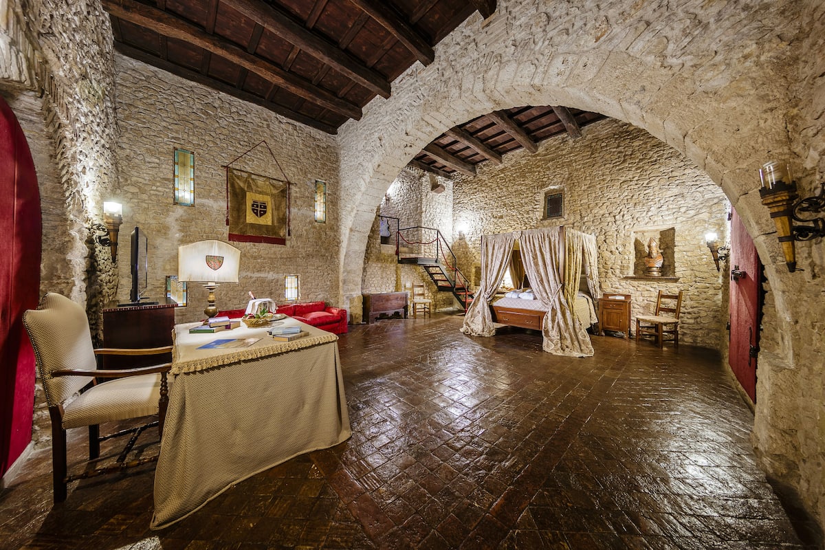 Una habitación rústica con paredes de piedra, vigas de madera, una mesa con libros, una silla y una cama con dosel.
