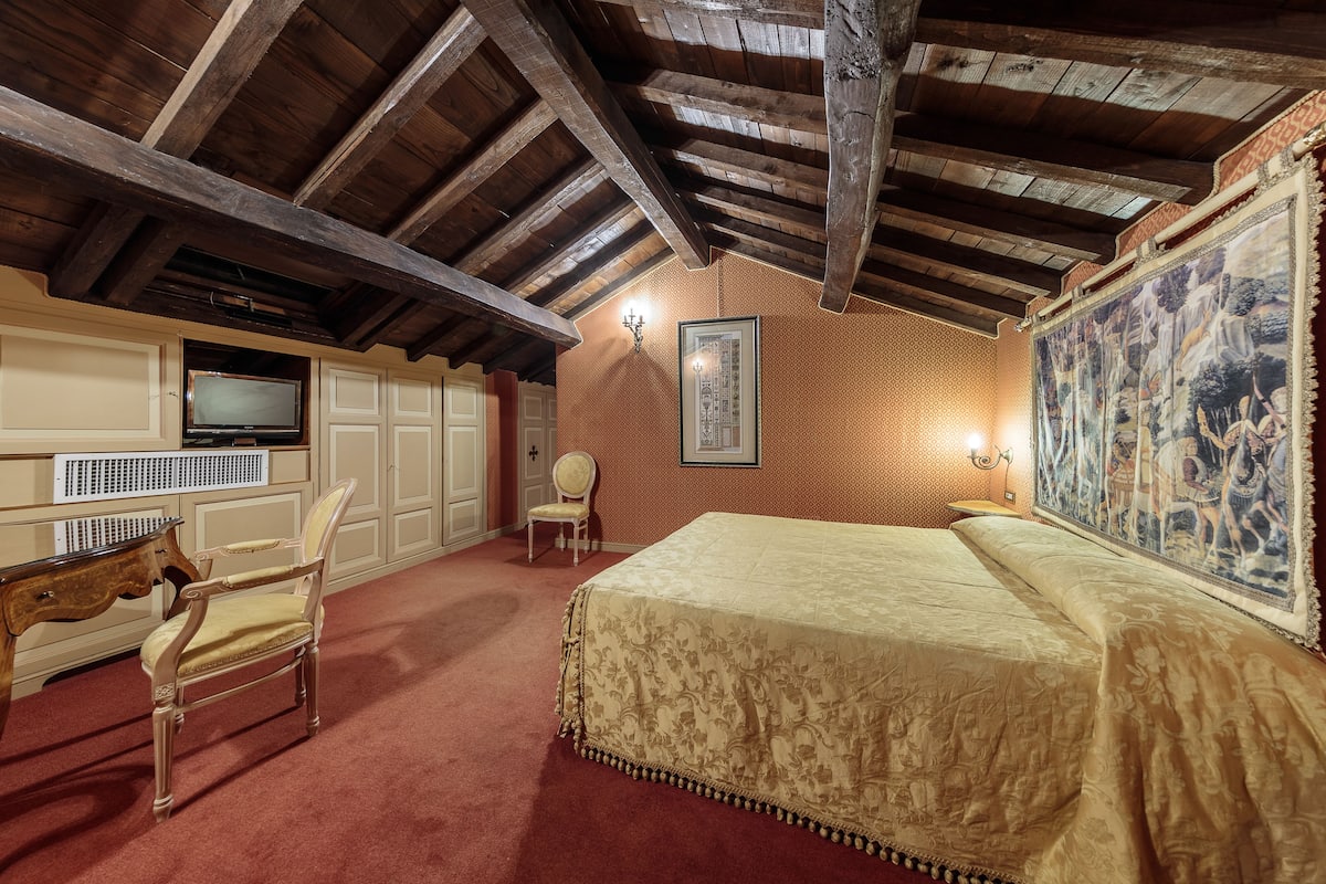Un dormitorio con una cama grande, vigas de madera en el techo, un televisor, una silla y un cuadro en la pared.