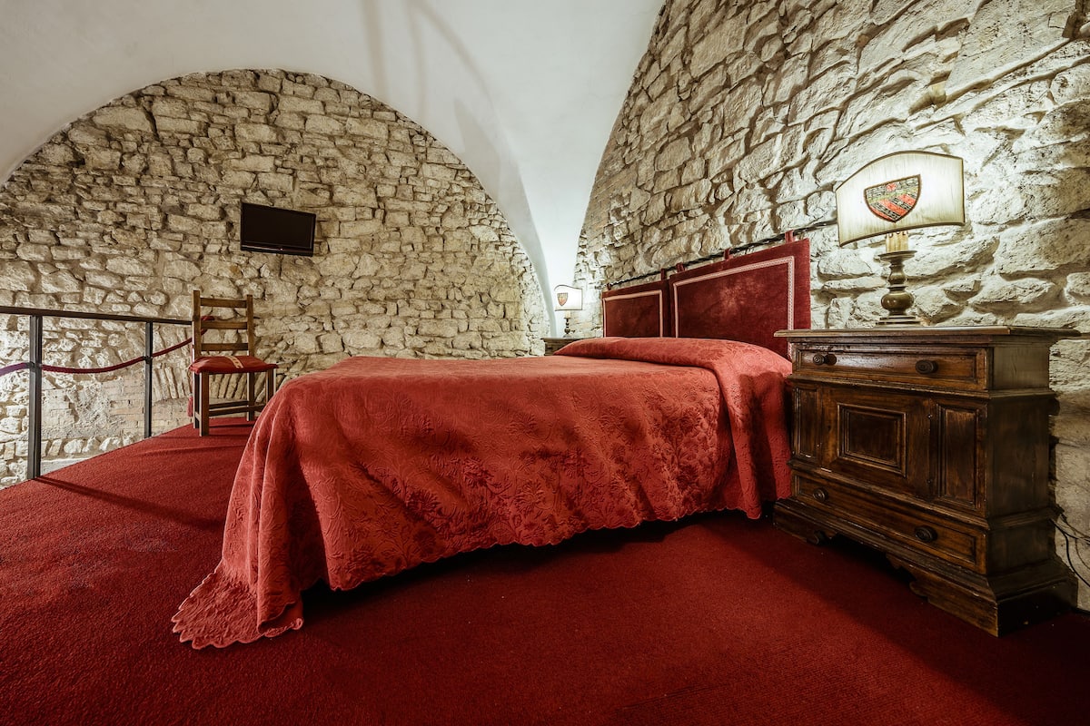 Una habitación con pared de piedra, una cama con colcha roja, un aparador de madera y una lámpara con pantalla en forma de escudo.