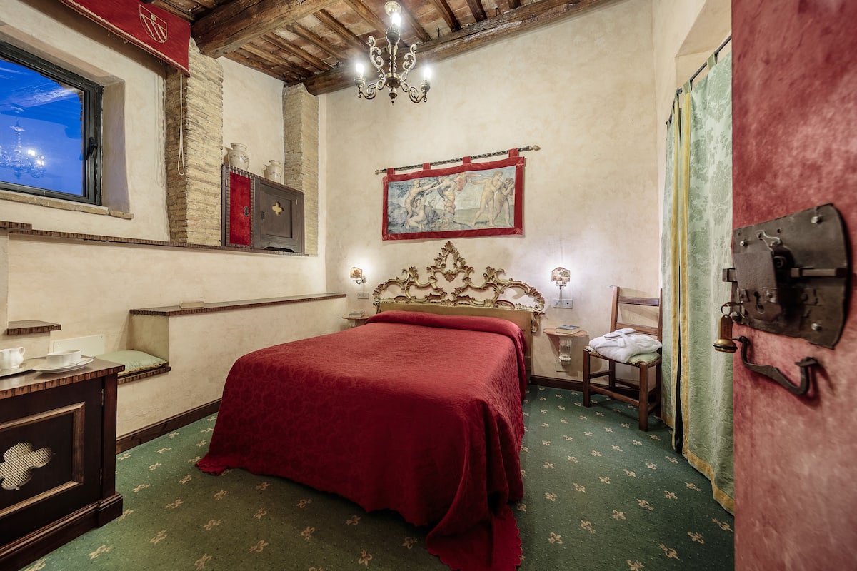 Una habitación con cama, un armario de madera, una ventana, una araña y un cuadro en la pared.