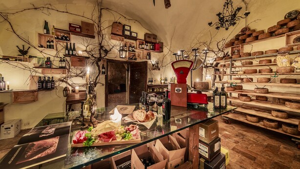 A rustic interior with a glass table displaying food and wine, surrounded by shelves of bottles and cheese. - Castello Orsini Hotel (Nerola)