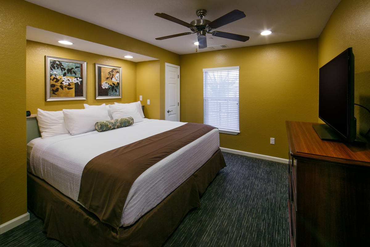 A hotel room with a large bed, a ceiling fan, a flat-screen TV, and two framed floral paintings on the wall.