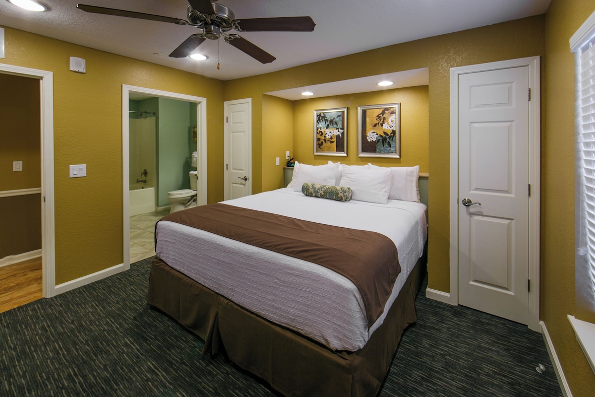A hotel room with a bed, a ceiling fan, a bathroom with a shower, and two framed pictures on the wall.