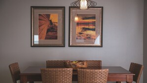 A dining area with a wooden table and wicker chairs, framed artwork on the wall, and a woven basket on the table.