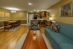 A living room with a blue sofa, wooden coffee table, and patterned armchair. There is a dining area with wooden chairs and a table, and a kitchen with wooden cabinets and a refrigerator.