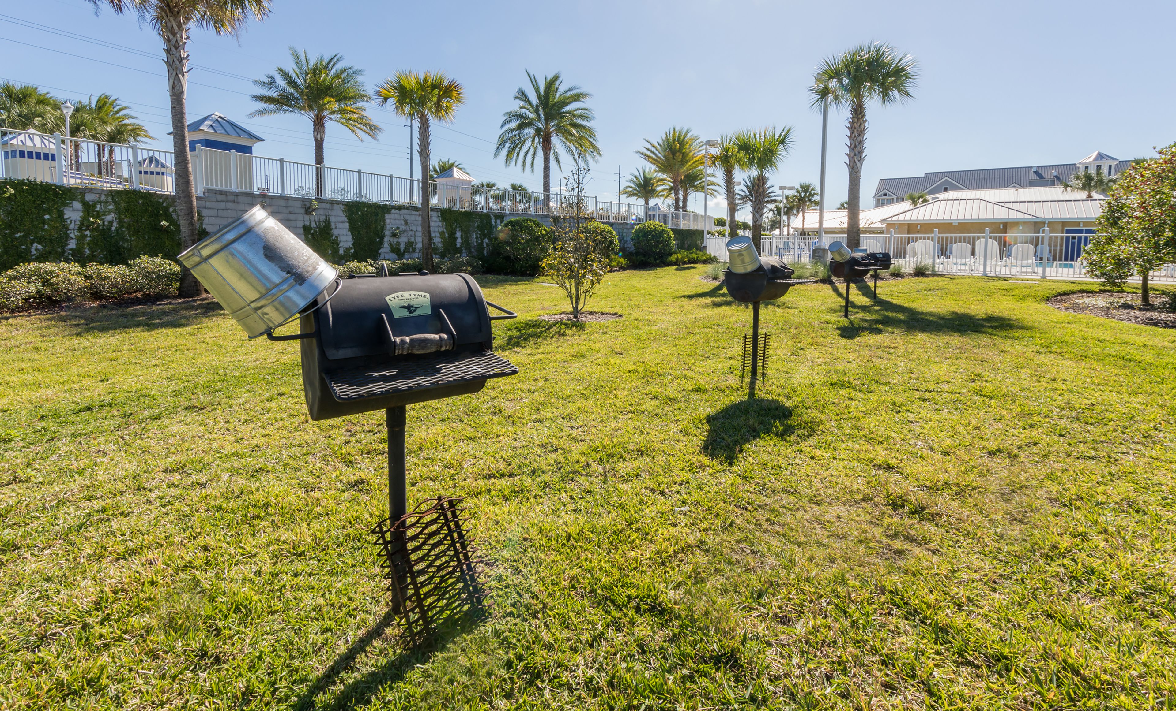 A grassy outdoor area with barbecue grills, palm trees, and a white fence.