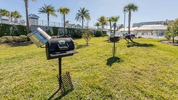 A grassy outdoor area with barbecue grills, palm trees, and a white fence.