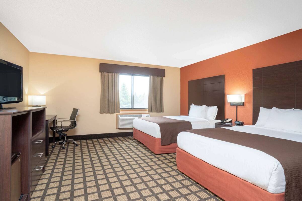 A hotel room with two beds, a desk with a TV, a chair, a window with curtains, and an air conditioning unit.