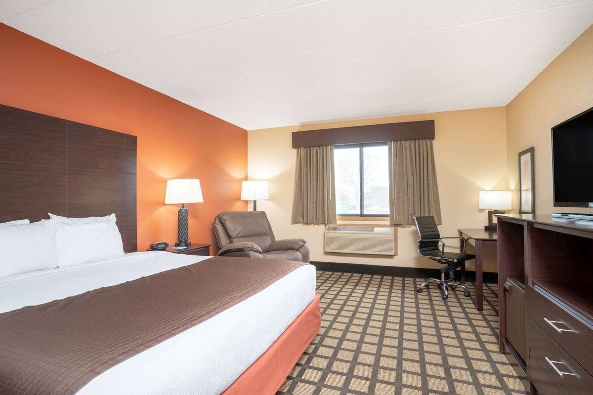 A hotel room with a bed, two chairs, a desk, a television, and a window with curtains.