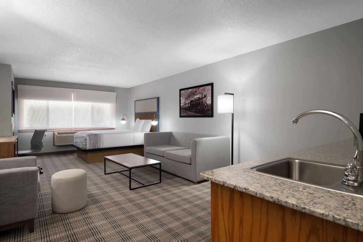 A modern hotel room with a sofa, ottoman, coffee table, and a kitchen area with a sink and countertop.