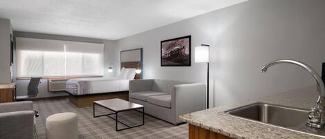 Una habitación de hotel moderna con sofá, reposapiés, mesa de centro y una zona de cocina con fregadero y encimera.