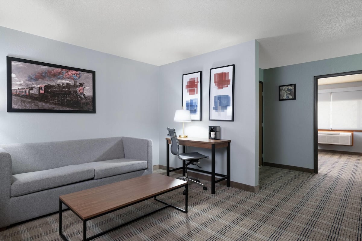 A hotel room with a grey sofa, a wooden coffee table, a desk with a chair, and framed artwork on the walls.