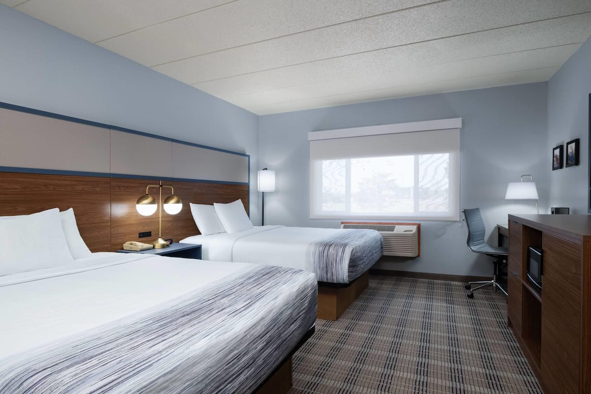 A hotel room with two beds, a desk, a chair, a lamp, and a window.
