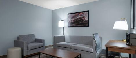 A hotel room with a grey sofa, a small round ottoman, a wooden coffee table, a desk with a lamp, and a framed picture on the wall.