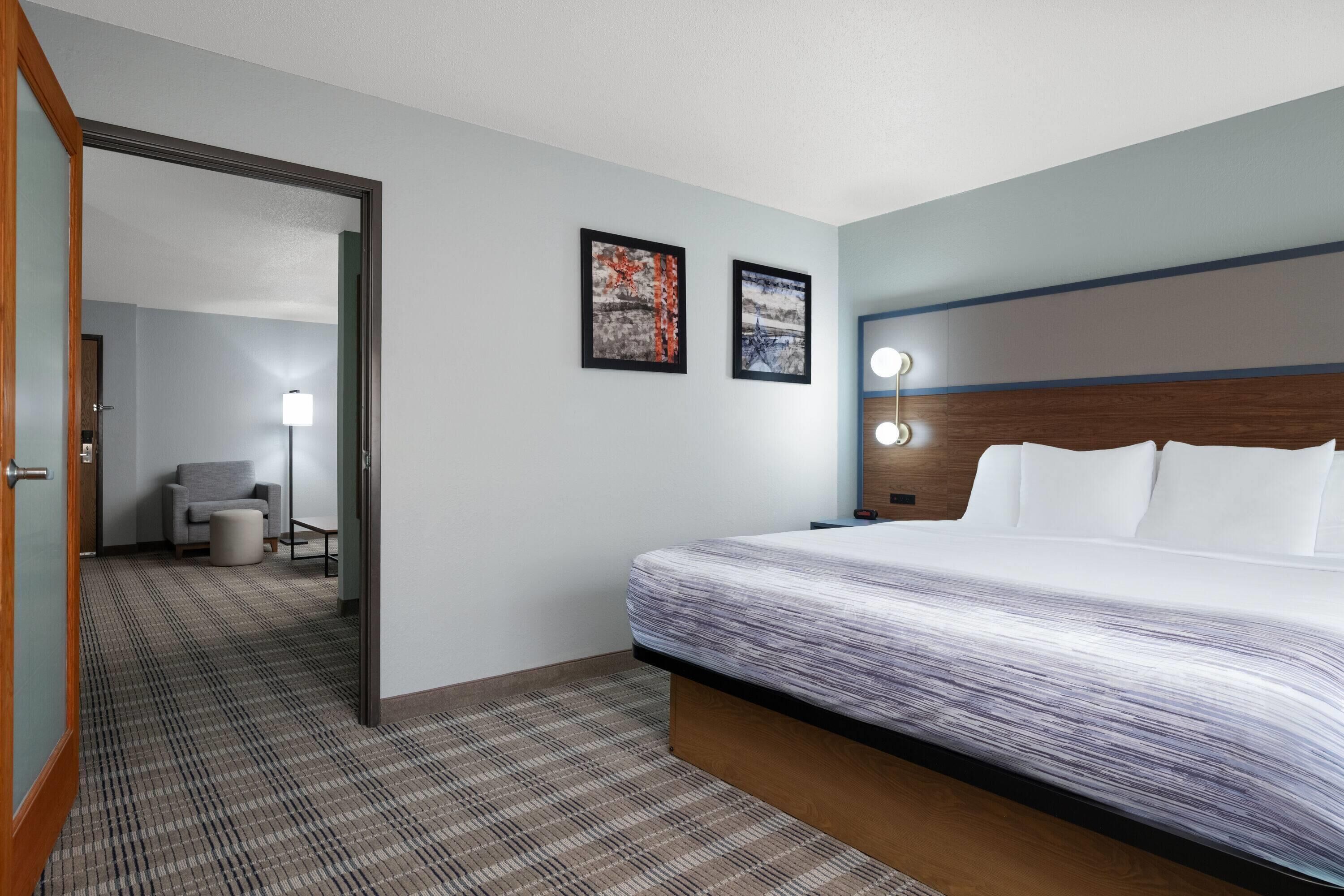 A hotel room with a large bed, a wooden headboard, two framed pictures on the wall, a small seating area with a chair and table, and a carpeted floor.