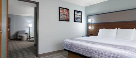A hotel room with a large bed, a wooden headboard, two framed pictures on the wall, a small seating area with a chair and table, and a carpeted floor.