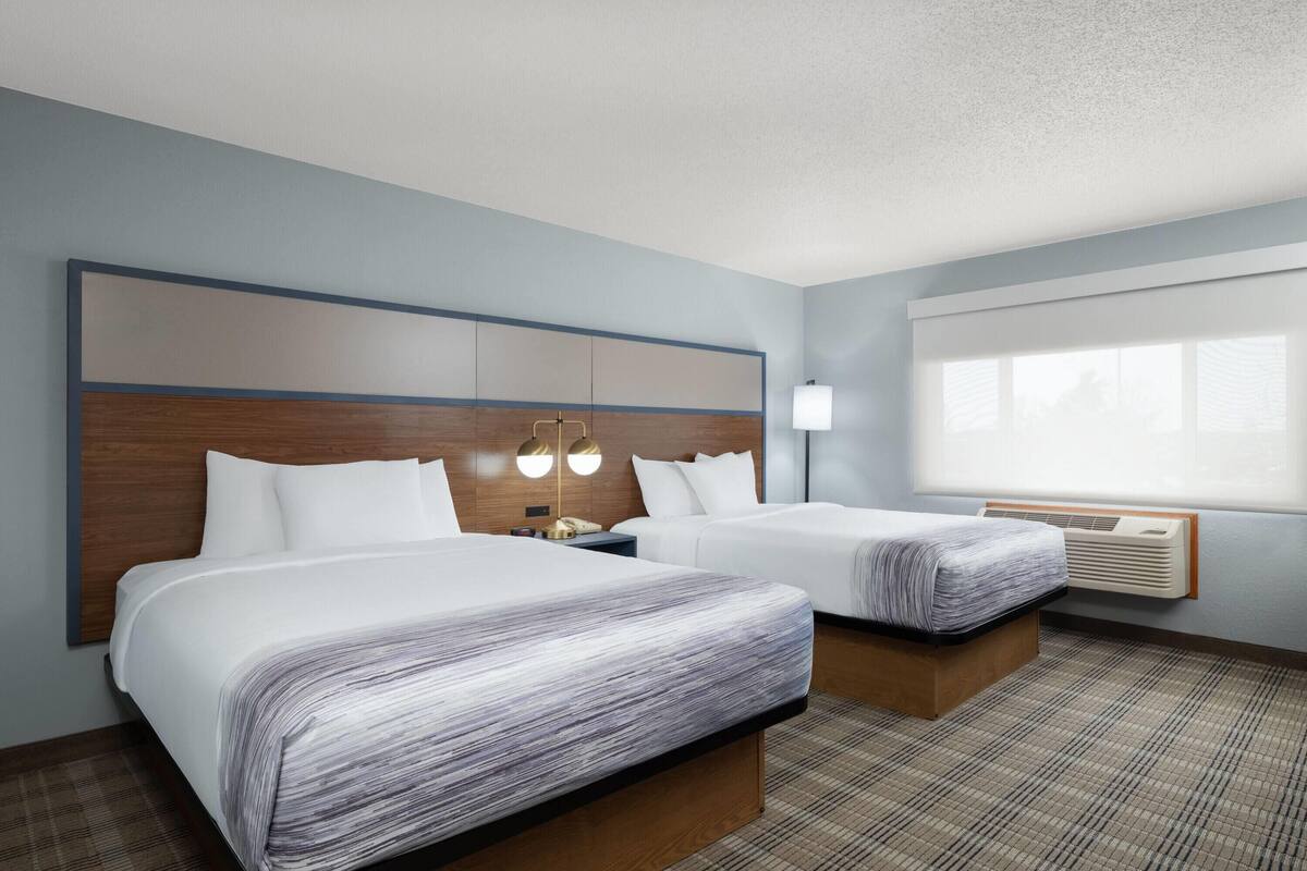 A hotel room with two beds, a large window, a wall-mounted air conditioner, and a headboard with built-in lighting.