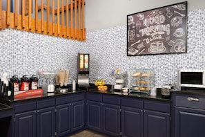 A kitchen with a black countertop, blue cabinets, a coffee station, and a 'Good Morning Welcome' sign. - AmericInn by Wyndham Baldwin (Baldwin)