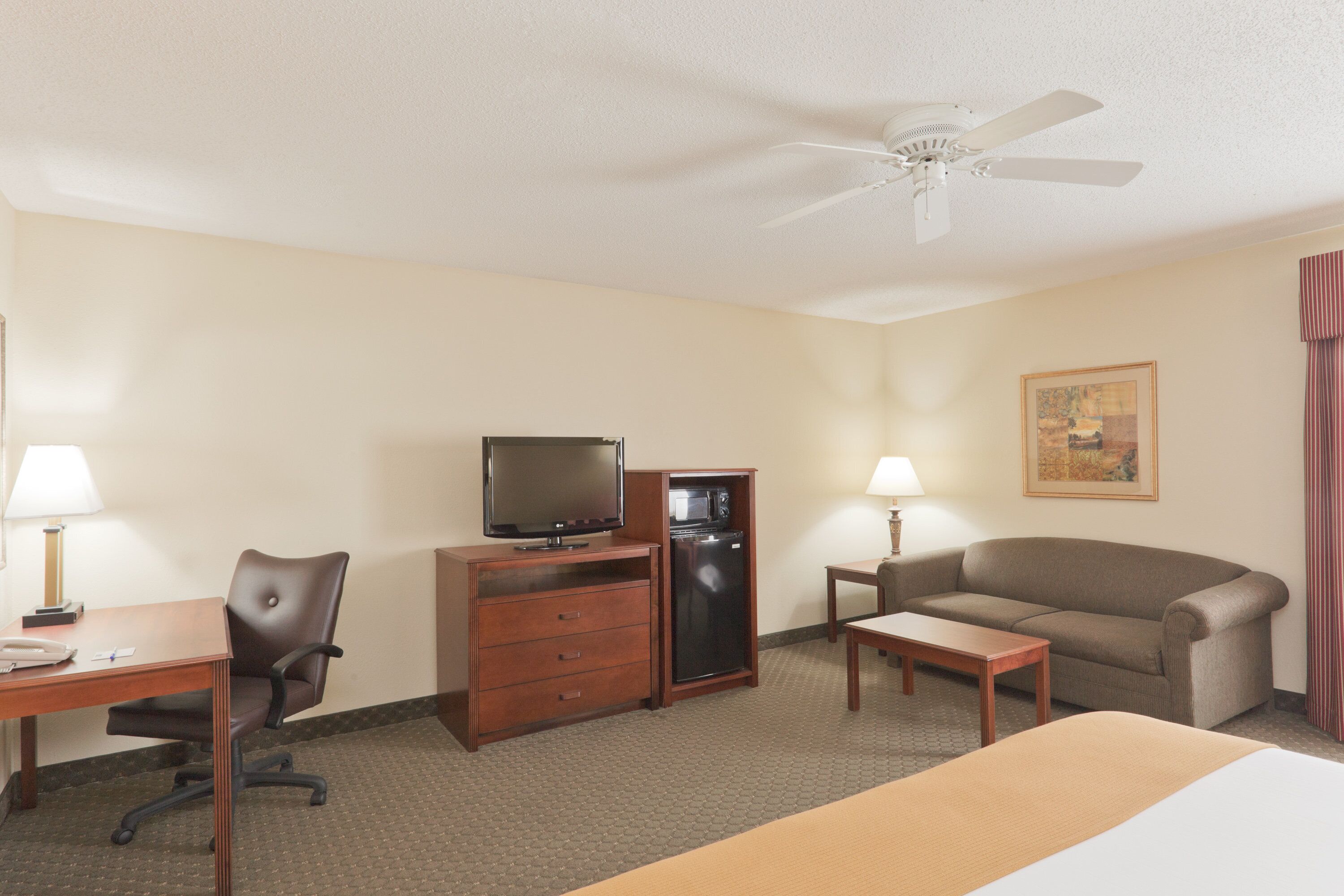 A hotel room with a bed, a desk with a chair, a television, a refrigerator, a sofa, and a ceiling fan.