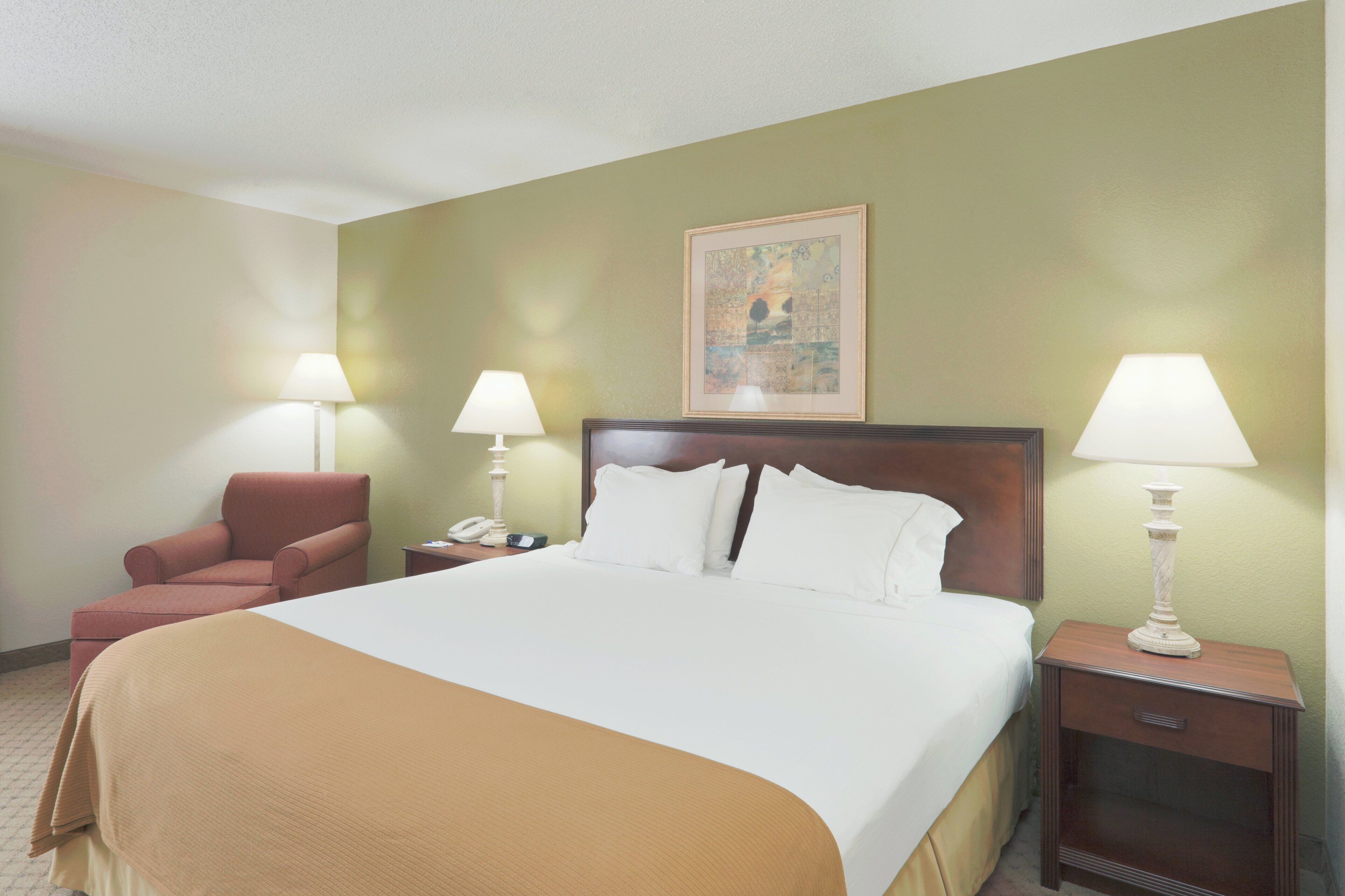 A hotel room with a large bed, two bedside lamps, a chair, a nightstand with a phone, and a painting on the wall.