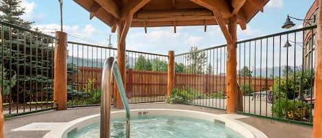 Un bain à remous sous une pergola en bois, dotée d’une rambarde en métal, entouré d’une clôture et de verdure.