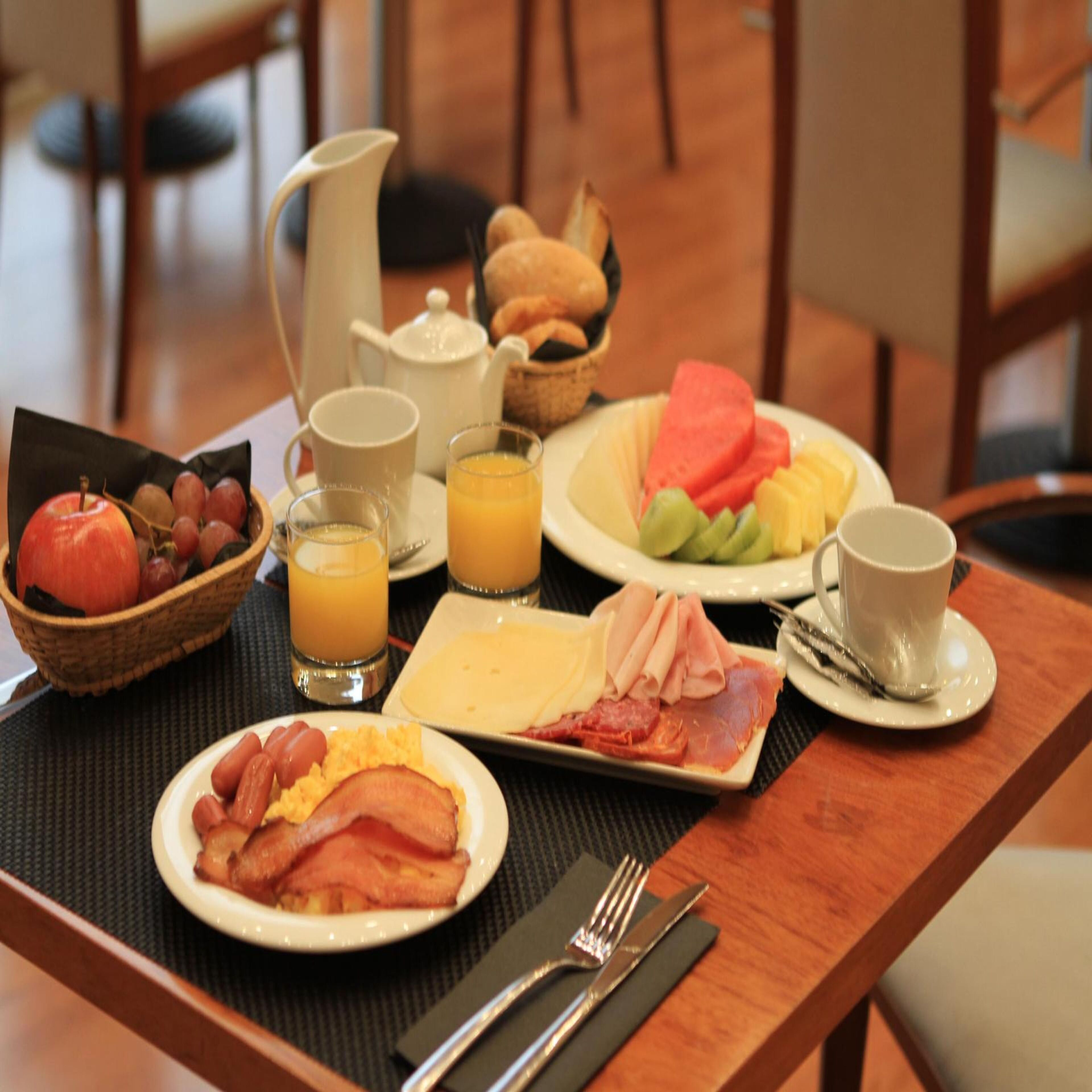 Een ontbijttafel met borden met eieren, spek, ham, kaas en fruit, samen met glazen sinaasappelsap en een koffiekop.