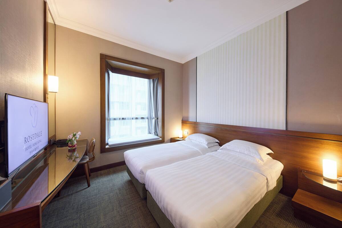 A hotel room with two beds, a desk, a TV, and a window with curtains.