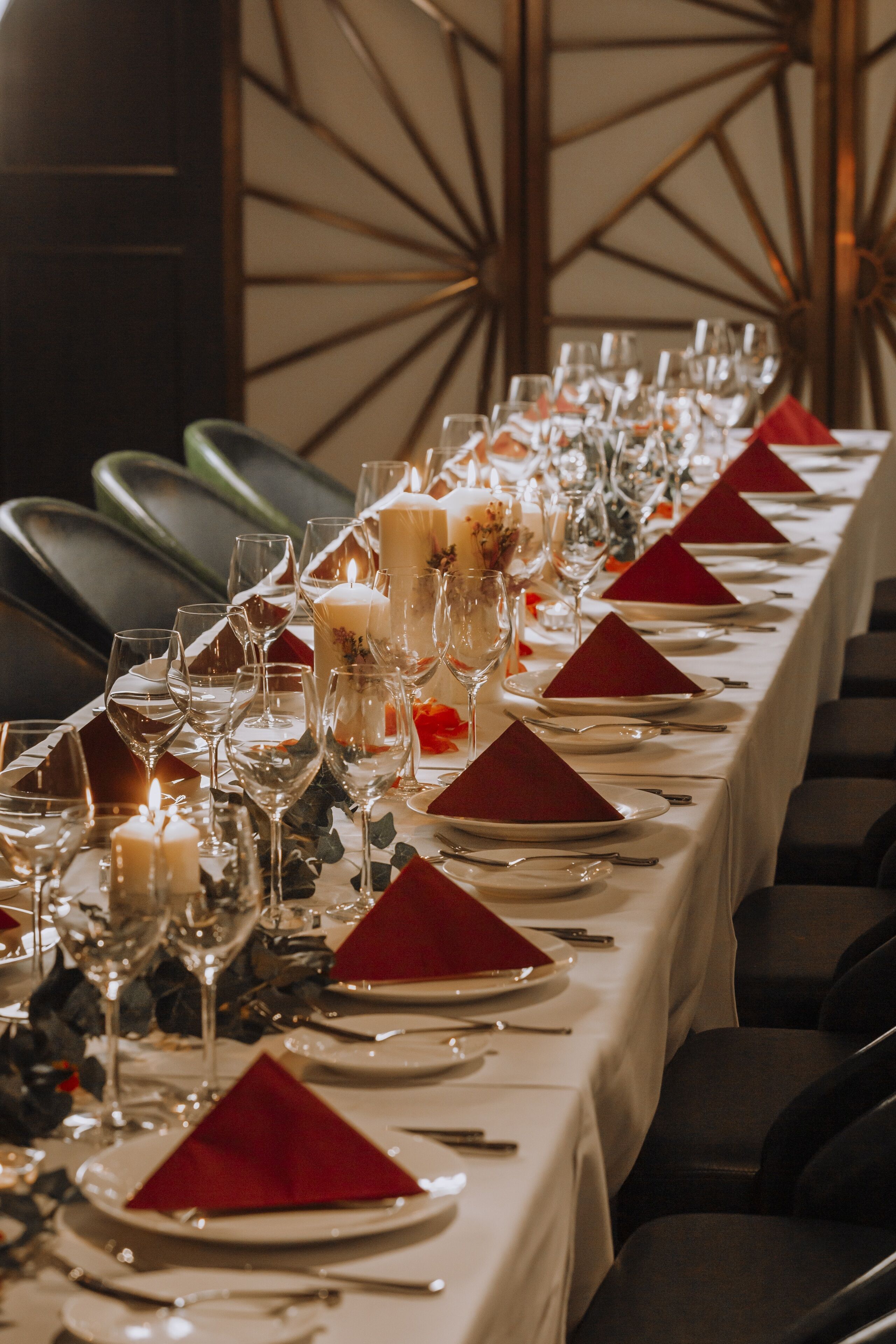 A long dining table set with red napkins, white plates, and clear glasses, featuring candles and floral decorations.