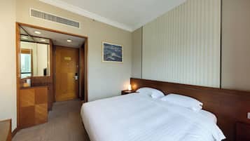 A hotel room with a large bed, a nightstand with a lamp, a wooden desk, a mirror, and a door leading to another room.