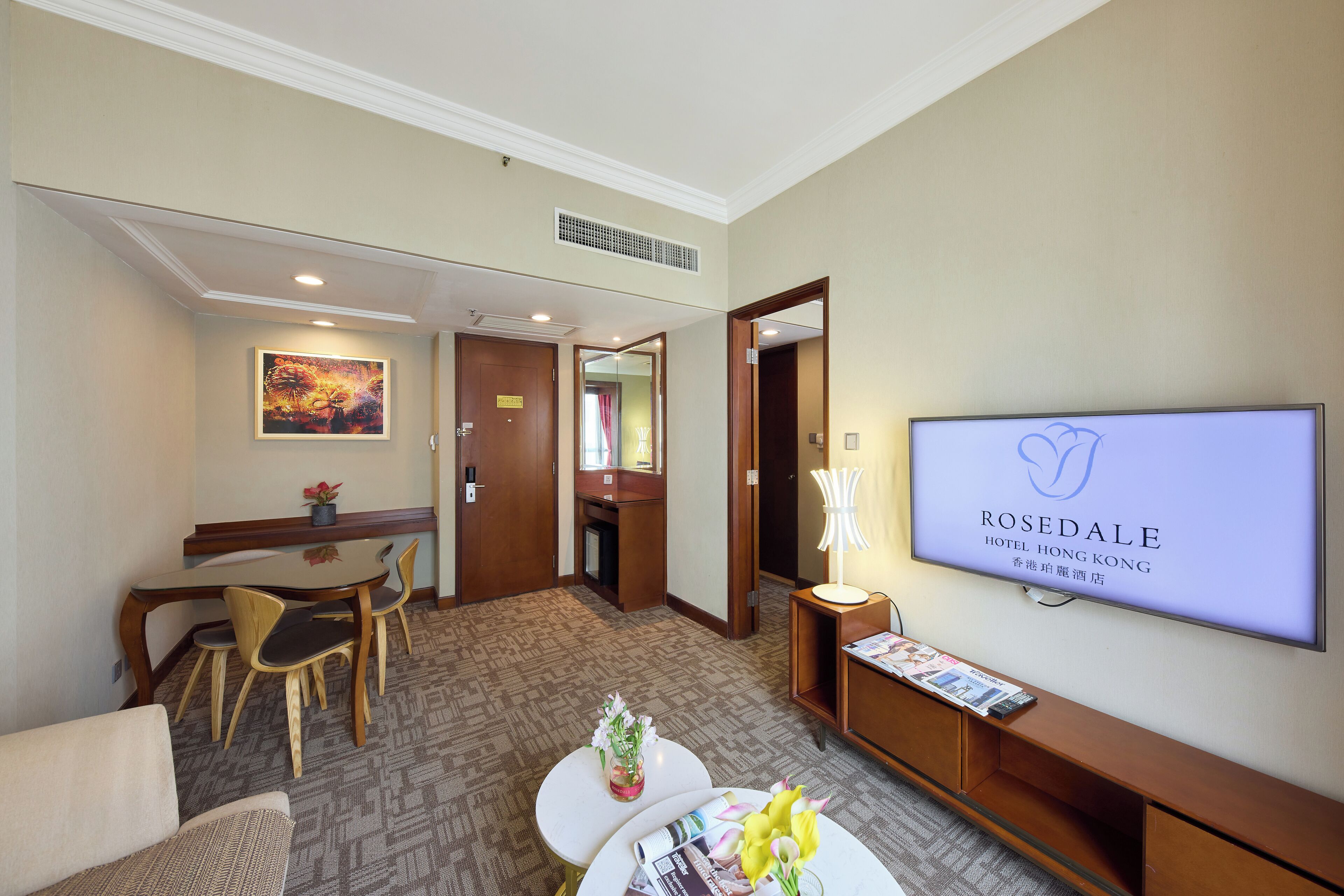A hotel lobby with a reception desk, a television displaying the hotel name, a dining area with chairs and a table, and a small seating area with a sofa and a coffee table.