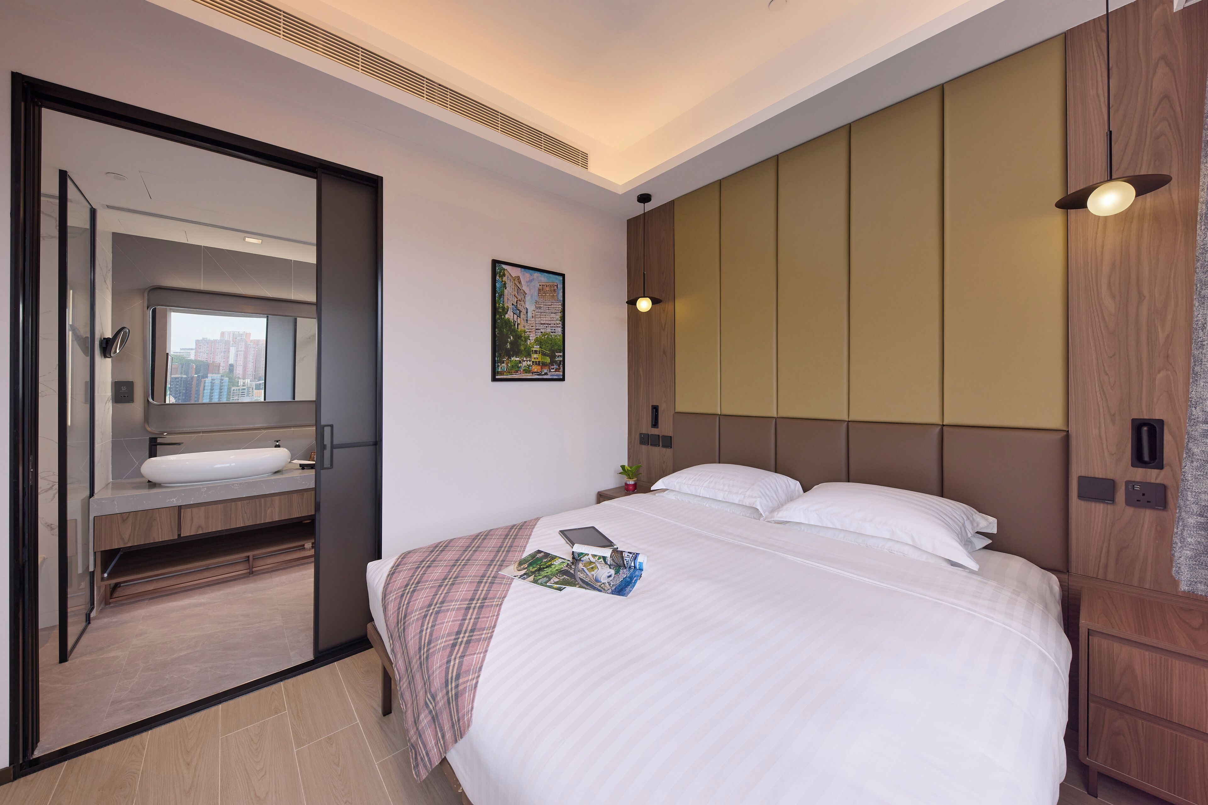 A hotel room with a bed, a bedside table, a nightstand, a mirror, a bathroom with a sink, and a cityscape view.