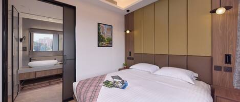 A hotel room with a bed, a bedside table, a nightstand, a mirror, a bathroom with a sink, and a cityscape view.