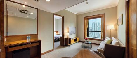 A hotel room with a wooden desk, a bed, a sofa, a small table with a lamp, and a large window with a city view.