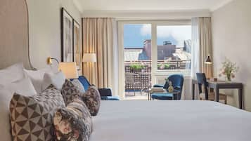 A hotel room with a large bed, a desk with a chair, a balcony with a view of buildings, and a window with curtains.