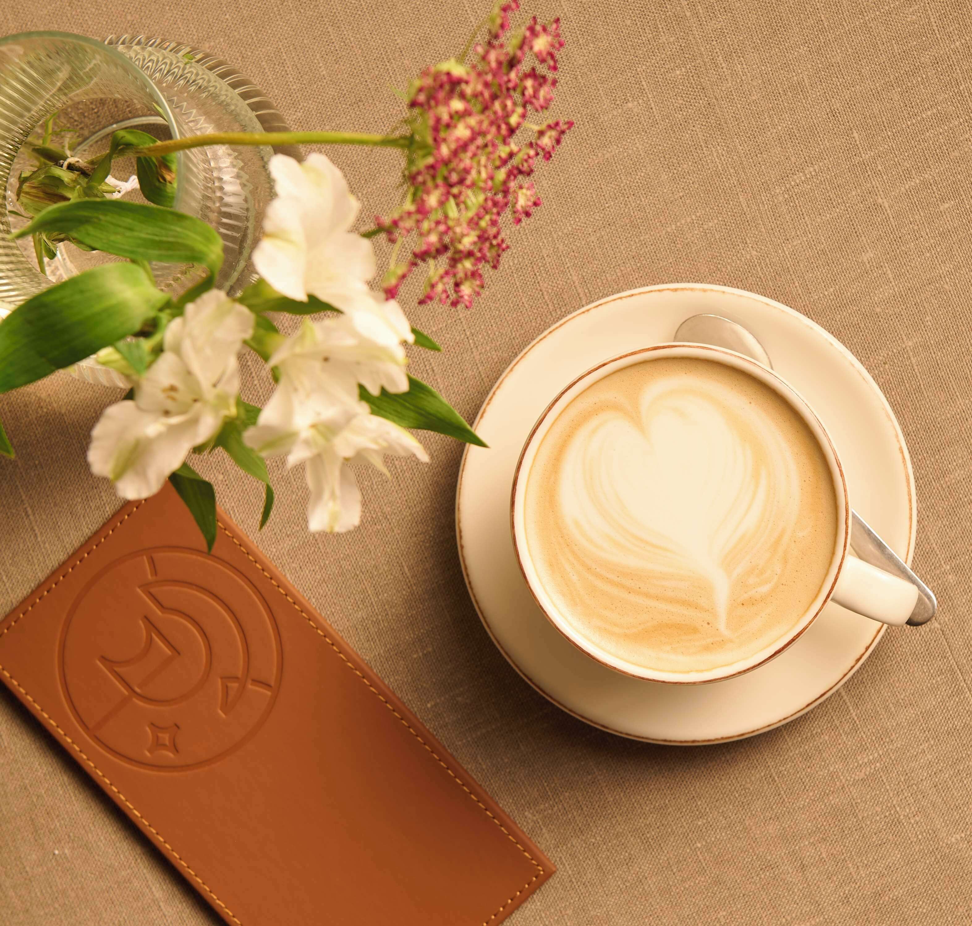 Een kop koffie met latte-art op een schotel, een glazen vaas met bloemen en een lederen portemonnee met een logo.