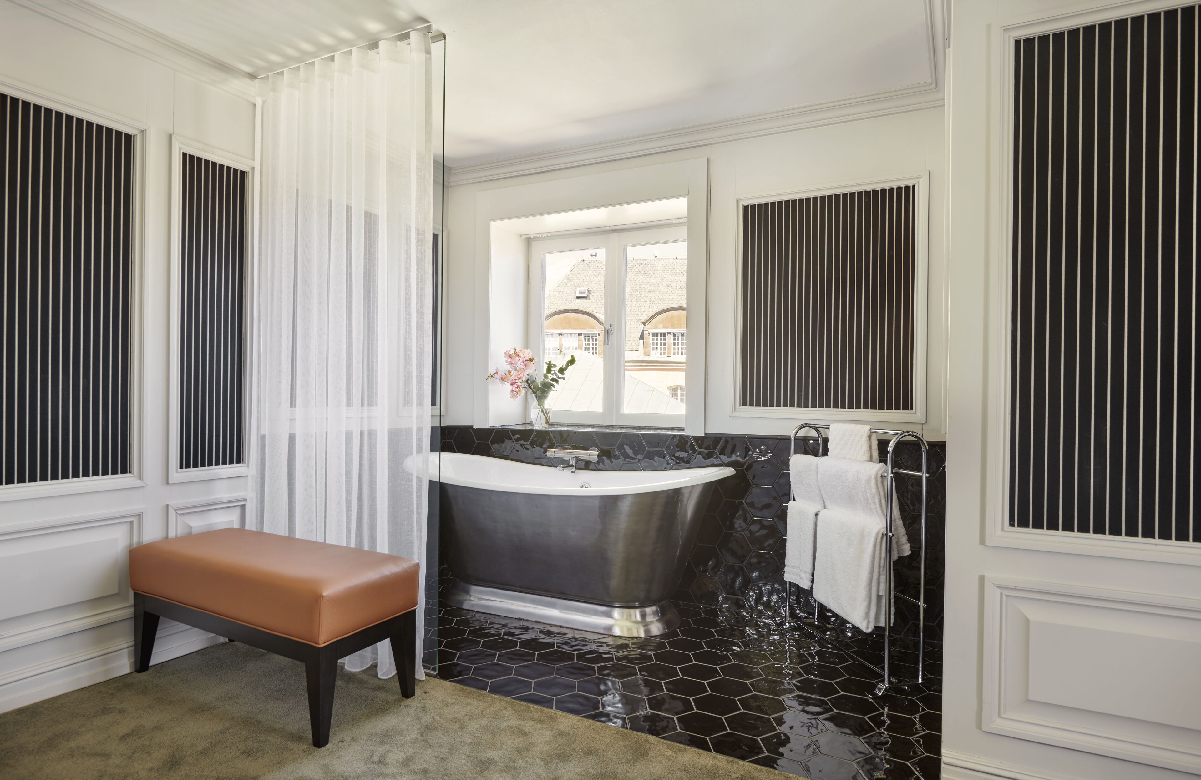 A bathroom with a freestanding bathtub, a bench, a window with curtains, and a view of buildings outside.