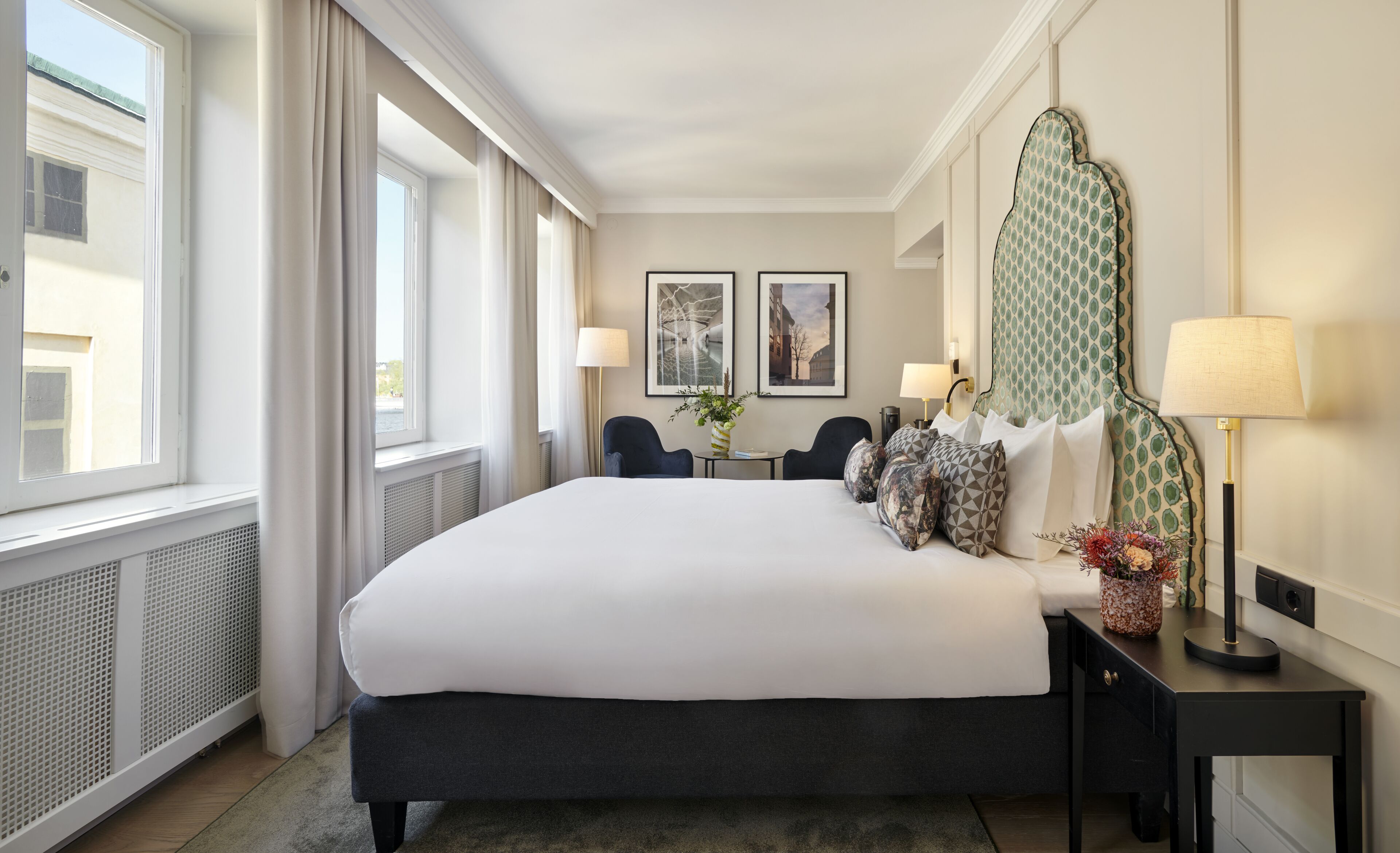 Quarto de hotel com uma cama grande, duas cadeiras, uma mesa com um vaso e duas fotografias emolduradas na parede.