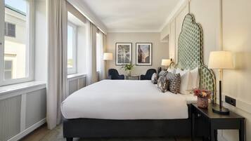 Quarto de hotel com uma cama grande, duas cadeiras, uma mesa com um vaso e duas fotografias emolduradas na parede.