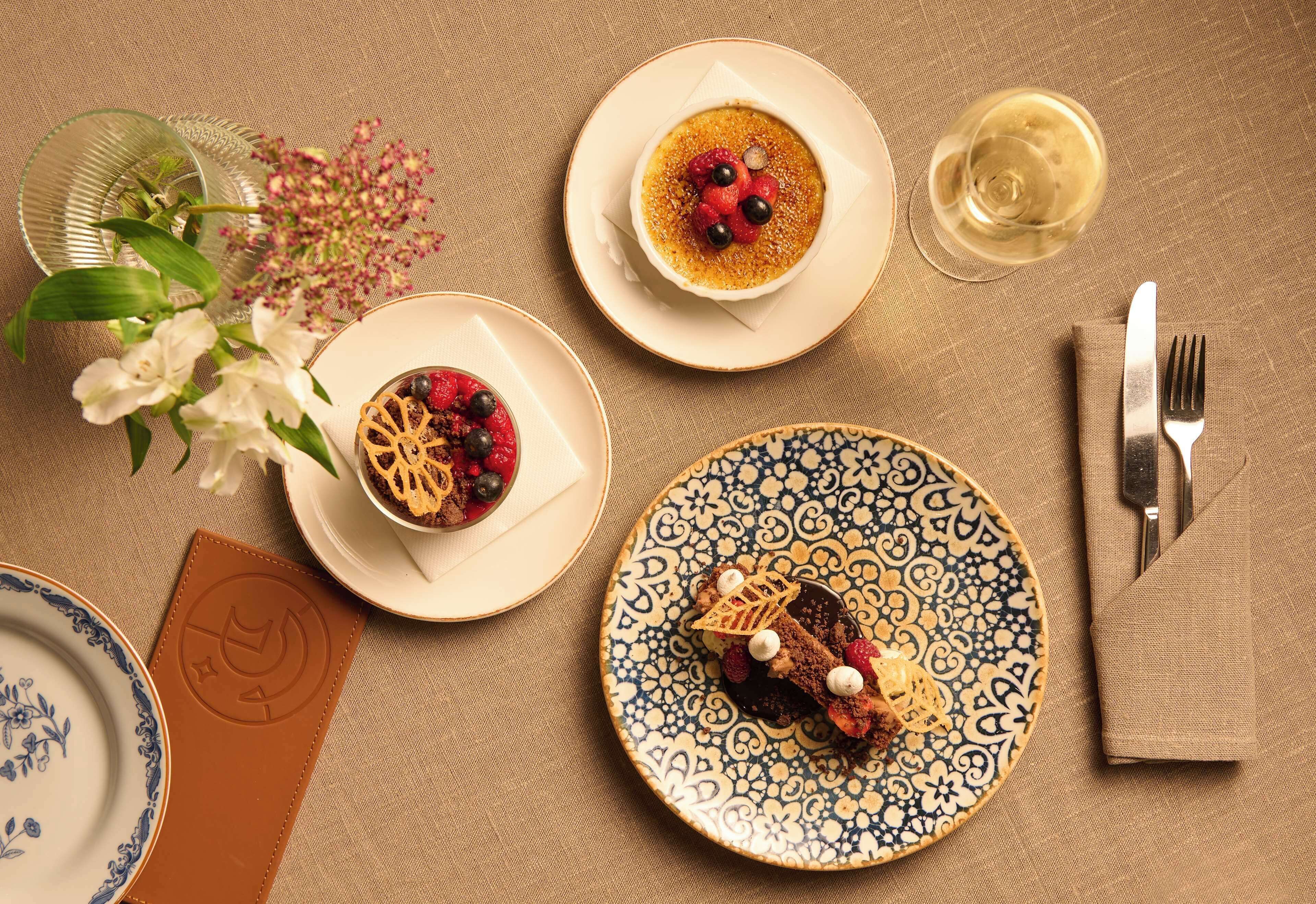 Una mesa dispuesta con platos de postres, una copa de vino blanco y un florero con flores.