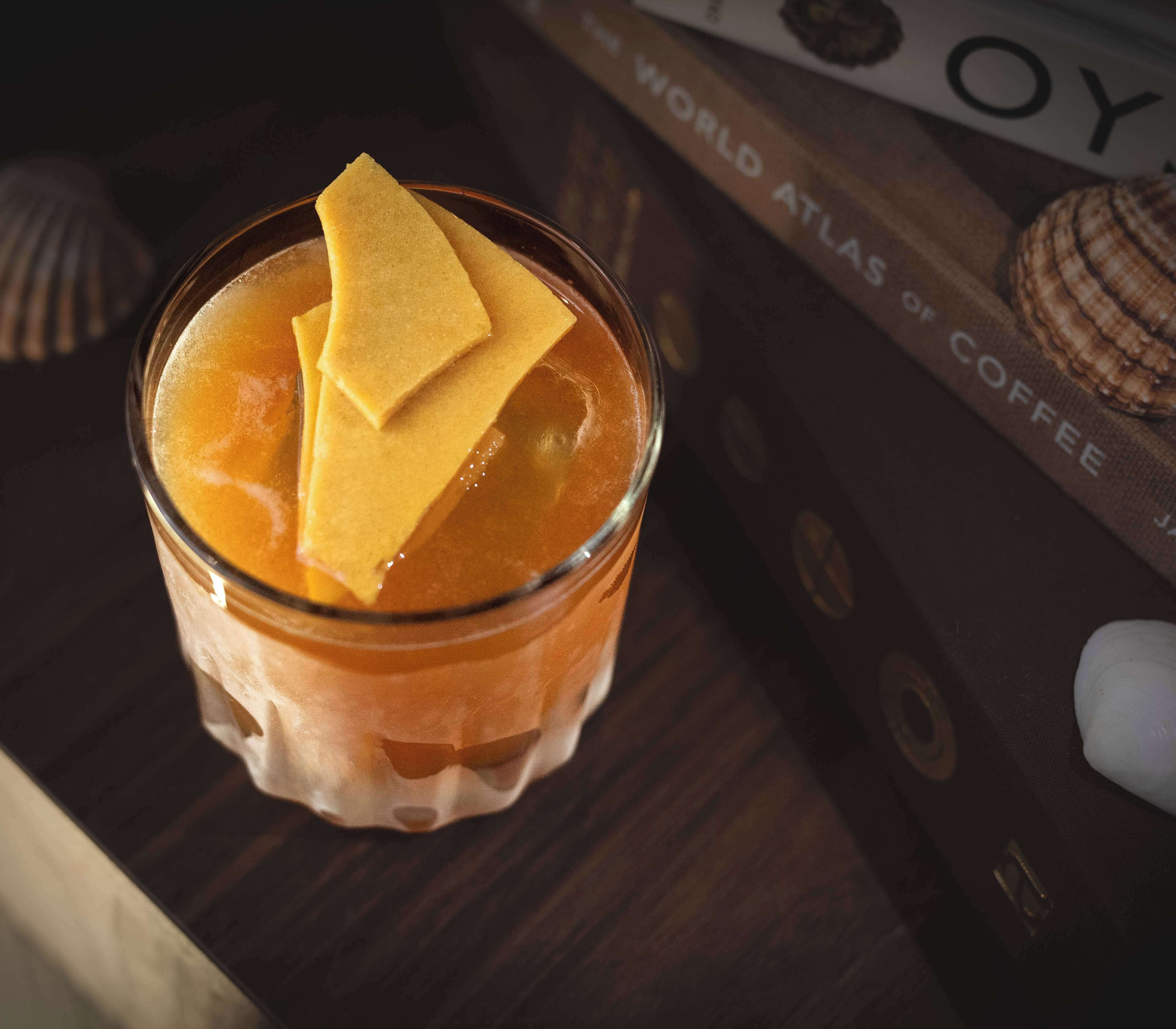 Een glas met een gelaagde drank en een vierkant versiering, geplaatst op een houten ondergrond, met een boek getiteld 'World Atlas of Coffee' op de achtergrond.