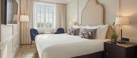 A hotel room with a large bed, two bedside lamps, a desk with a lamp, a chair, and a view of the city through the window.