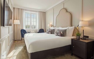 Un cuarto de hotel con una cama grande, dos lámparas de noche, un escritorio con lámpara, una silla y vista a la ciudad por la ventana.