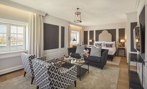 Ein modernes Hotelzimmer mit einem großen Bett, einer Couch und zwei Sesseln. Durch das Fenster bietet sich ein Blick auf die Stadt.