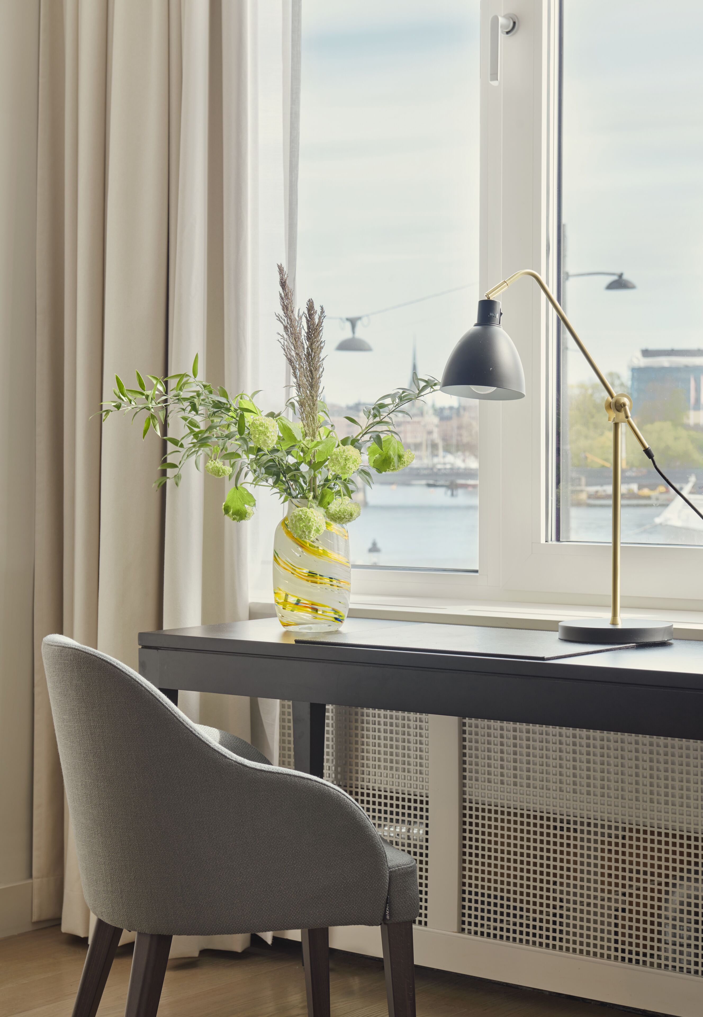 A desk with a vase of green plants, a lamp, and a chair in front of a window overlooking a river.