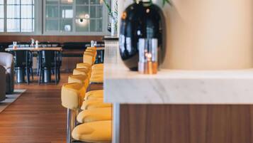 Een restaurantinterieur met een lange toonbank, gele barkrukken en een grote vaas met groen.
